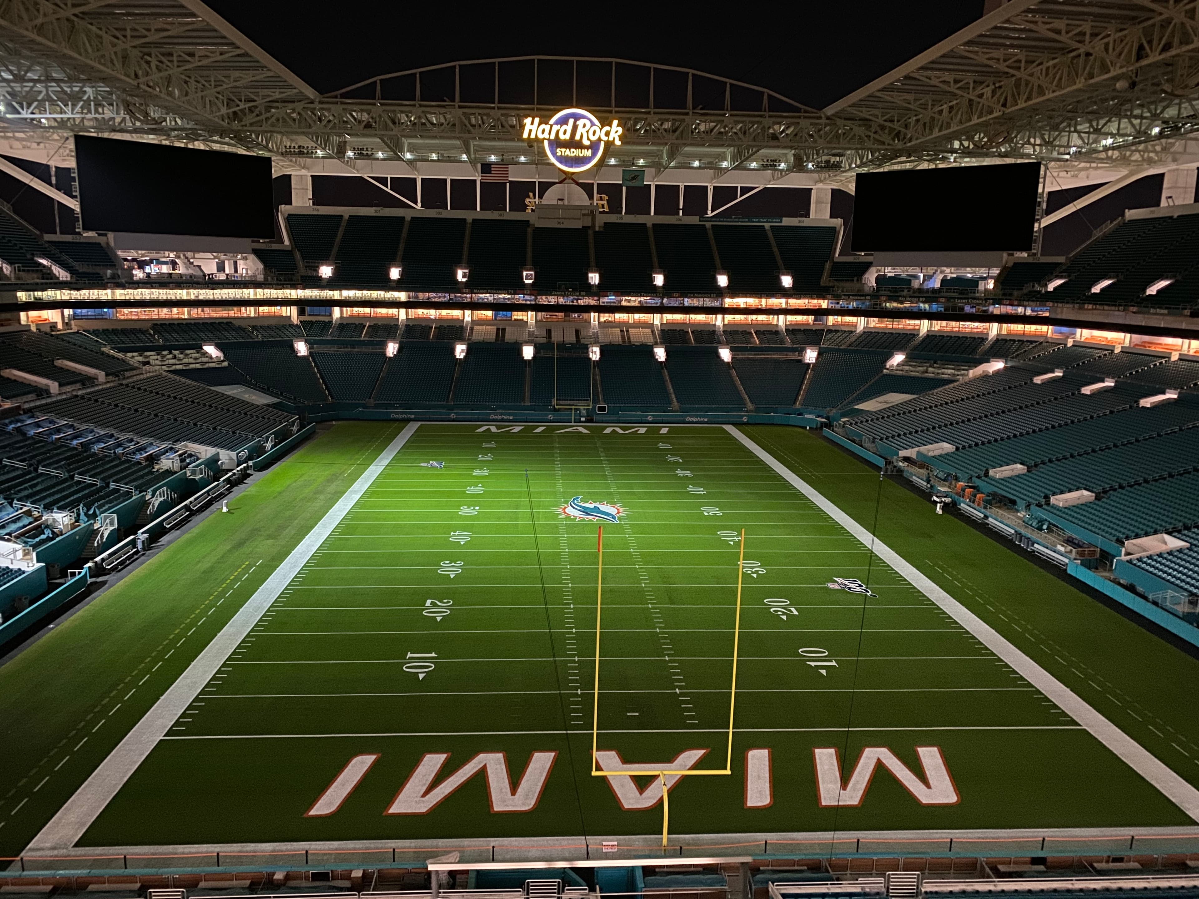 Hard Rock Stadium at night — the empty bowl with the field lit, the Miami Dolphins logo at midfield, and the glowing Hard Rock Stadium sign above